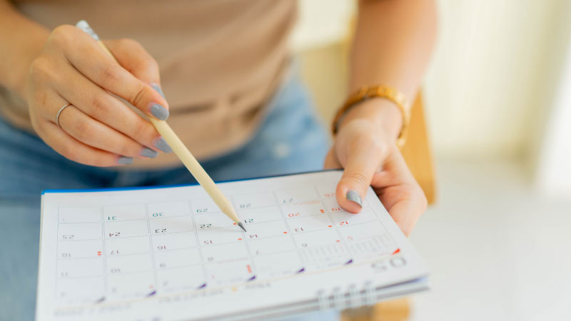 A woman with painted nails holds a pencil and the calendar. She points at the calendar with the pencil. 