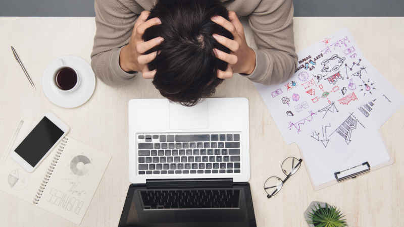 frustrated employee holding his head in his hands due to stress at workplace, surrounded by laptop, cellphone, coffee cup and reports