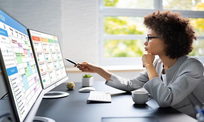 Project manager checking and updating tasks and milestones progress planning with scheduling interface on a desktop computer with two screens
