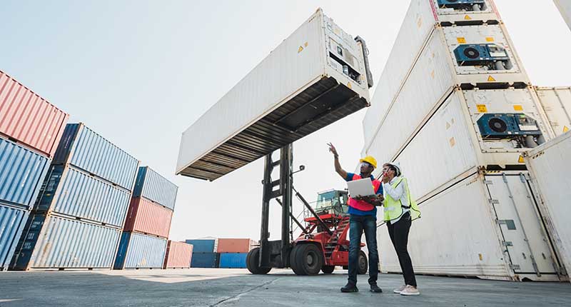 Black foreman and a woman worker checking a container cargo harbor holding laptop and using walkie talkie