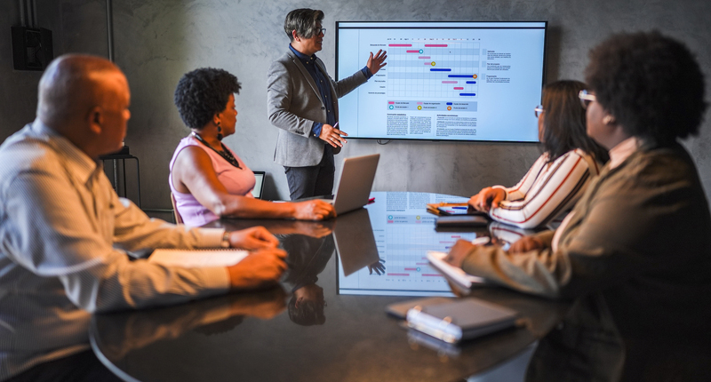 A professional explains a chart on a screen about winning in a consulting business in a meeting while other four professionals seated across a table follow the presentation intently